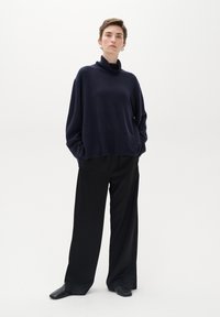 Jūras spēku pelēka turtlenecka džemperis ar brīvu piegriezumu, apvienots ar platām melnām biksēm; modelis stāv ar rokām kabatās uz vienkārša baltā fona.