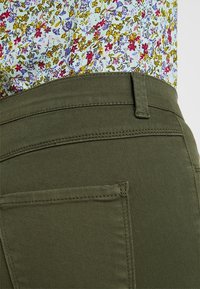 Olivgrüne Hosen verfügen über eine nahtlose Textur und eine Seitentasche. Das Oberteil hat ein leichtes Blumenmuster mit lila, gelben und roten Akzenten.