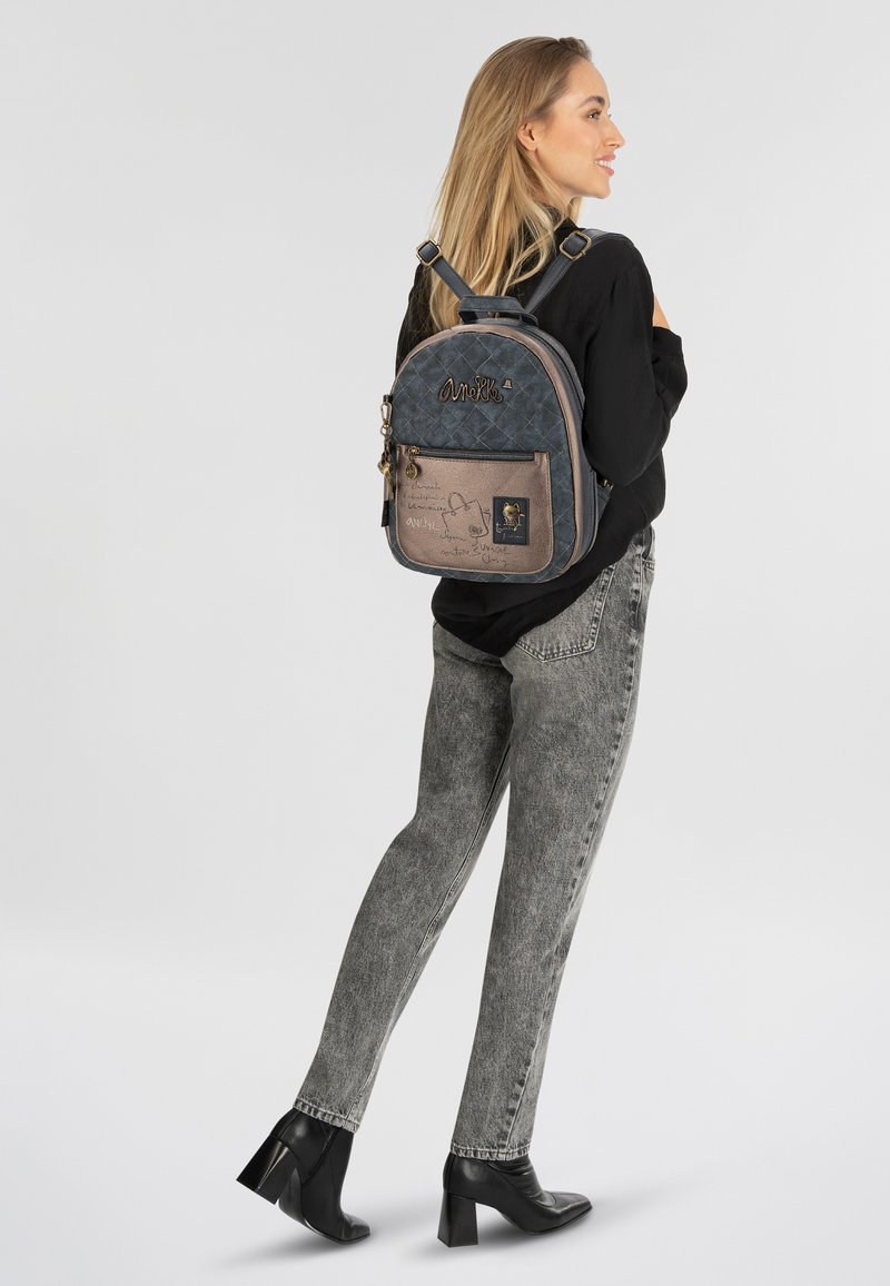 Blau- und grauer, gesteppten Rucksack mit einer bronzefarbenen Fronttasche, ausgestattet mit einem Reißverschluss, dekorativer Schrift und einem Logo-Patch.