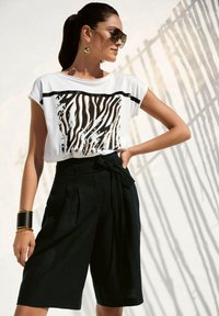 Weißes T-Shirt mit schwarzen und orangen Zebra-Streifen, kombiniert mit hoch taillierten schwarzen Culottes mit einem Stoffbindegürtel. Die Accessoires umfassen auffällige Ohrringe.