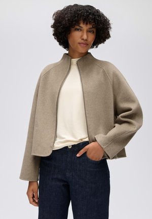 Donna con capelli ricci che indossa una giacca beige con cerniera sopra una maglietta bianca e jeans blu scuro, con una mano in tasca, in piedi davanti a uno sfondo uniforme.