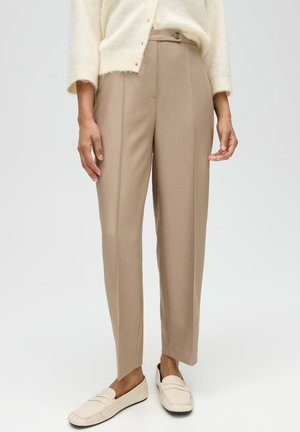 Vrouw draagt beige op maat gemaakte broek, crèmekleurige trui en crèmekleurige instap loafers, staand tegen een effen lichte achtergrond.