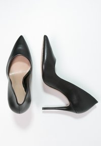 Paire de chaussures à talons hauts noires à bout pointu, l’une vue du dessus et l’autre de côté, sur un fond blanc.
