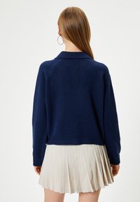 Marineblauer Strickcardigan mit einem kragenartigen Ausschnitt und geripptem Saum. Das Modell trägt einen plissierten beigen Rock. Rückansicht, die Schulternaht ist im Raglan-Stil.