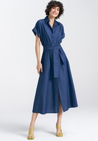 Niebieska sukienka koszulowa z dżinsu z wcięciem w talii, krótkimi podwiniętymi rękawami, zapięciem na guziki z przodu i bocznym rozporkiem, zestawiona z zielonymi sandałami na paski.