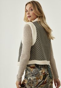 Chaleco de patrón negro y crema con un collar y dobladillo de texturizado de piel sintética, llevado sobre un suéter ligero y una falda floral colorida.