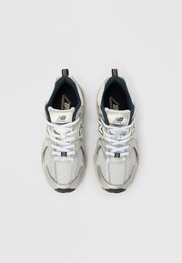 Šedé a bílé sportovní tenisky kombinují síťované a syntetické materiály, mají polstrovanou podrážku, tkaničky a poutko pro snadné nazouvání. Na jazyku je výrazné logo.