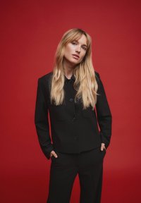 Sort skræddersyet jakke med knaplukning foran, to forlommer og lange ærmer, kombineret med matchende bukser mod en rød baggrund.