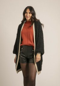 Cardigan noir en peluche avec des finitions beiges, porté sur un pull en laine rouge et un short en cuir noir taille haute, avec des boutons décoratifs.