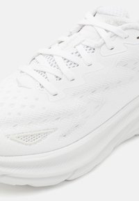 Zapatilla deportiva blanca con una parte superior de malla texturizada, cordones planos y suela acolchada con branding sutil y acentos reflectantes.