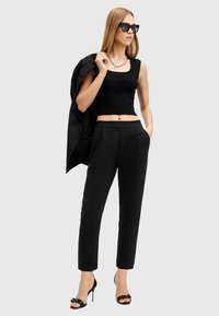 Top corto nero abbinato a pantaloni neri sartoriali e sandali con tacco alto. La modella tiene in mano un blazer nero, indossando occhiali da sole neri oversize.