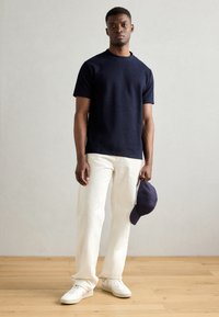 Marine strukturert t-skjorte, hvite vide bukser, hvite sneaker og en marineblå caps holdt i hånden, stående på et tregulv mot en lys vegg.
