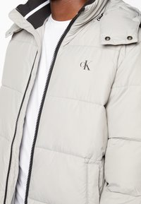 Chaqueta acolchada en gris claro con cremallera negra y cuello con botón a presión, llevada sobre una camisa blanca, con el logo "cK" en el pecho.