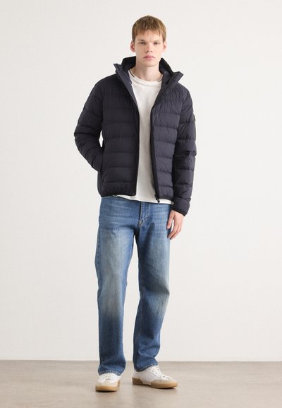 Doudoune marine avec des coutures horizontales, fermeture éclair à l'avant et col montant, portée sur un t-shirt blanc et un jean bleu, associée à des baskets blanches.