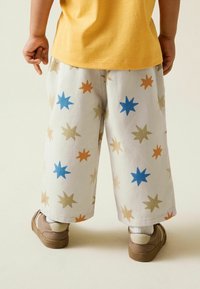 Weiße Hose mit Sternmustern in Blau, Orange und Beige. Kombiniert mit einem gelben Hemd und braunen Sneakers, mit einer lässigen Passform.