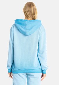 Hellblaue Kapuzenjacke aus weichem Stoff, mit Kapuze und gerippten Bündchen. "UNIVERSAL" in Weiß entlang des rechten Ärmels bedruckt.