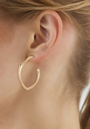 Gros plan sur l'oreille d'une personne montrant une boucle d'oreille en or brillante, de forme irrégulière, avec un design lisse et légèrement torsadé.