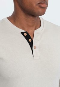 Mann in einem beigen Henley-Shirt mit drei hellbraunen Knöpfen und einem schwarzen Knopfdetails am Kragen.
