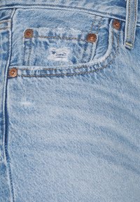Ausgeblichene blaue Denim-Jeans mit einer abgetragenen Tasche, ausgestattet mit kupfernen Nieten und sichtbaren Nähten. Strukturierter Stoff mit einem weichen, abgenutzten Aussehen.