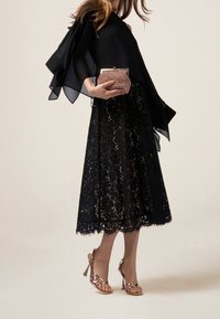 Jupe en dentelle noire avec ourlet festonné, haut en chiffon noir superposé avec bords volantés, escarpins à brides en métal rose doré, et pochette en tissu texturé.