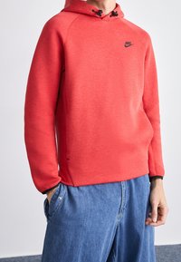 Man som bär en röd Nike-hoodie med svart logotyp och blå denimjeans, står med en hand i fickan mot en ljus bakgrund.