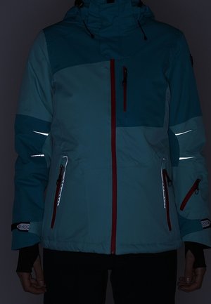 Blaue und türkisfarbene Winterjacke mit roten Reißverschlüssen und reflektierenden Streifen an Ärmeln und Taschen, getragen von einer Person, die mit den Händen nach unten steht.