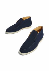 Zapatos azules marino de tipo slip-on con una textura de ante suave, puntera redondeada y suela de goma blanca en contraste. Diseño simple y minimalista.
