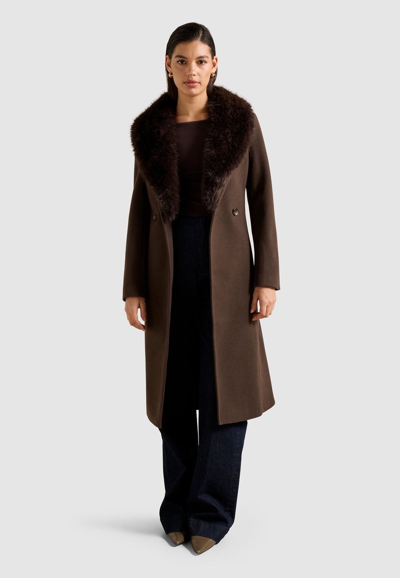 Donna che indossa un lungo cappotto marrone con un ampio colletto di pelliccia scura, top scuro, pantaloni a gamba larga e scarpe marroni appuntite, in piedi davanti a uno sfondo semplice.