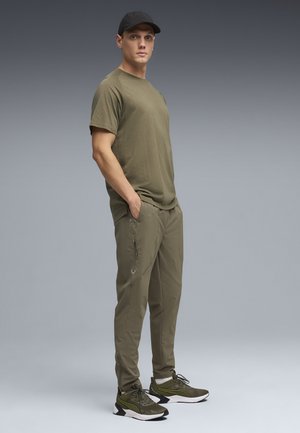 Młody mężczyzna stoi z rękami w kieszeniach, ubrany w oliwkowozielony t-shirt, pasujące spodnie dresowe, tenisówki i czarną czapkę na szarym tle.
