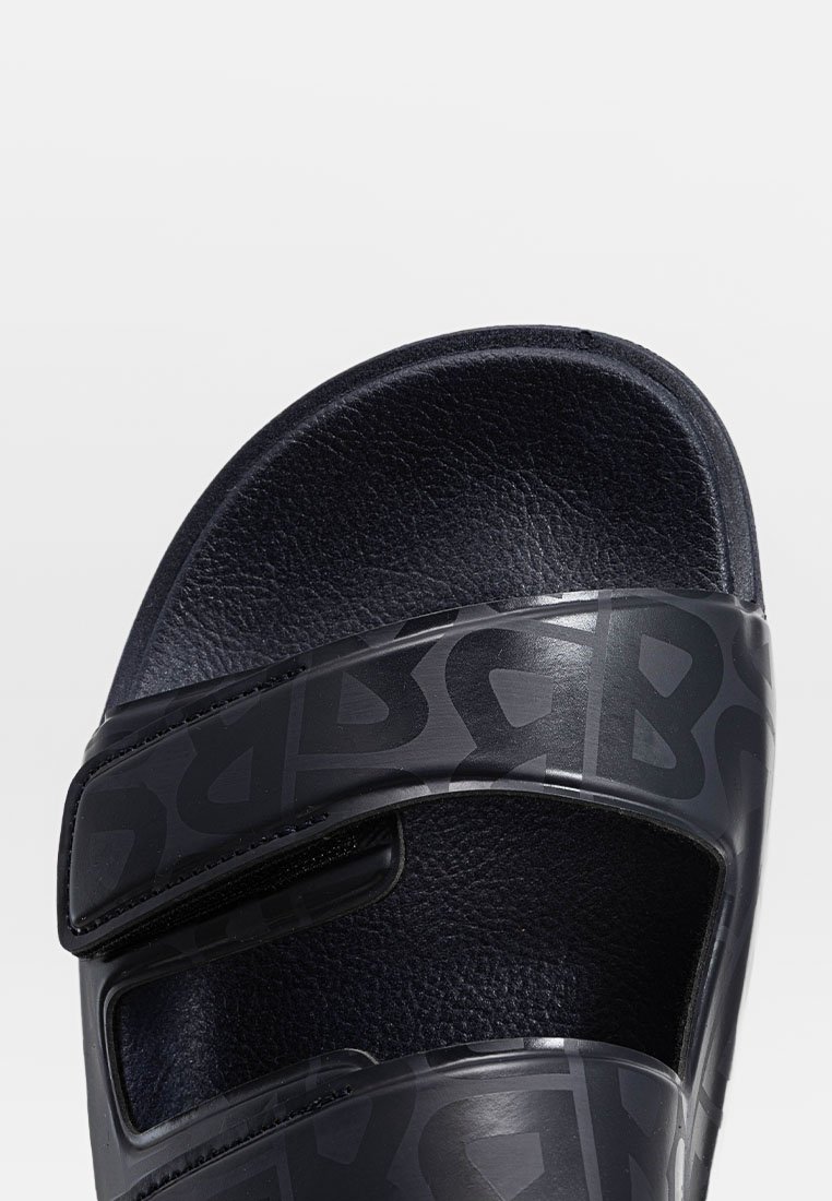 Schwarze Rutschsandale mit strukturiertem Obermaterial, verstellbaren Riemen und gepolsterter Innensohle. Auffälliges Markenmuster auf den Riemen.