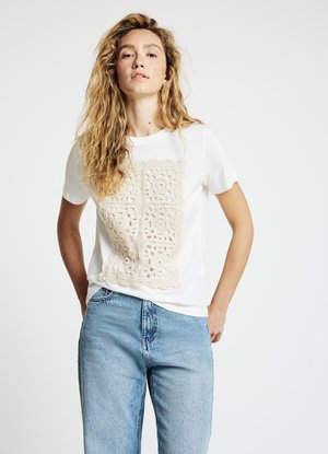 White T-shirt featuring a beige crochet design on the front. The T-shirt has short sleeves and a rounded neckline. Worn with light denim jeans.