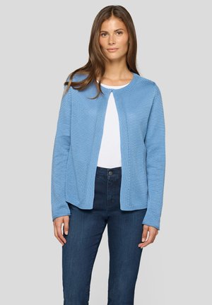 Frau, die eine hellblaue Strickjacke über einem weißen Oberteil und dunklen blauen Jeans trägt, steht vor einem einfachen hellgrauen Hintergrund.