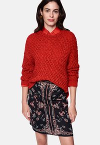 Red knitted sweater with a loose fit, featuring a textured design. Paired with a black skirt displaying floral patterns and stripes.