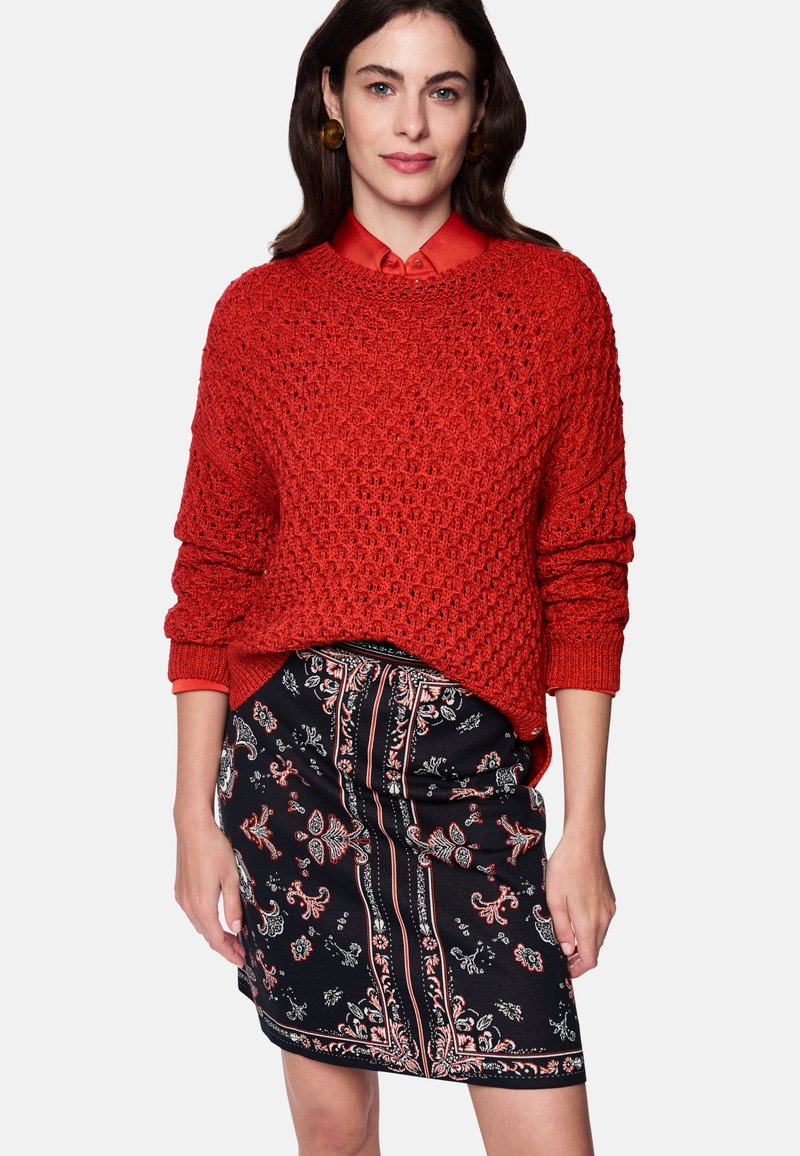 Red knitted sweater with a loose fit, featuring a textured design. Paired with a black skirt displaying floral patterns and stripes.