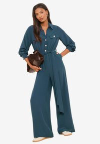 Türkisfarbener Overall mit langen Ärmeln, Knopfdetails und einem Taillengürtel. Verfügt über weite Hosenbeine und einen Kragen. Das Model hält eine braune Tasche.