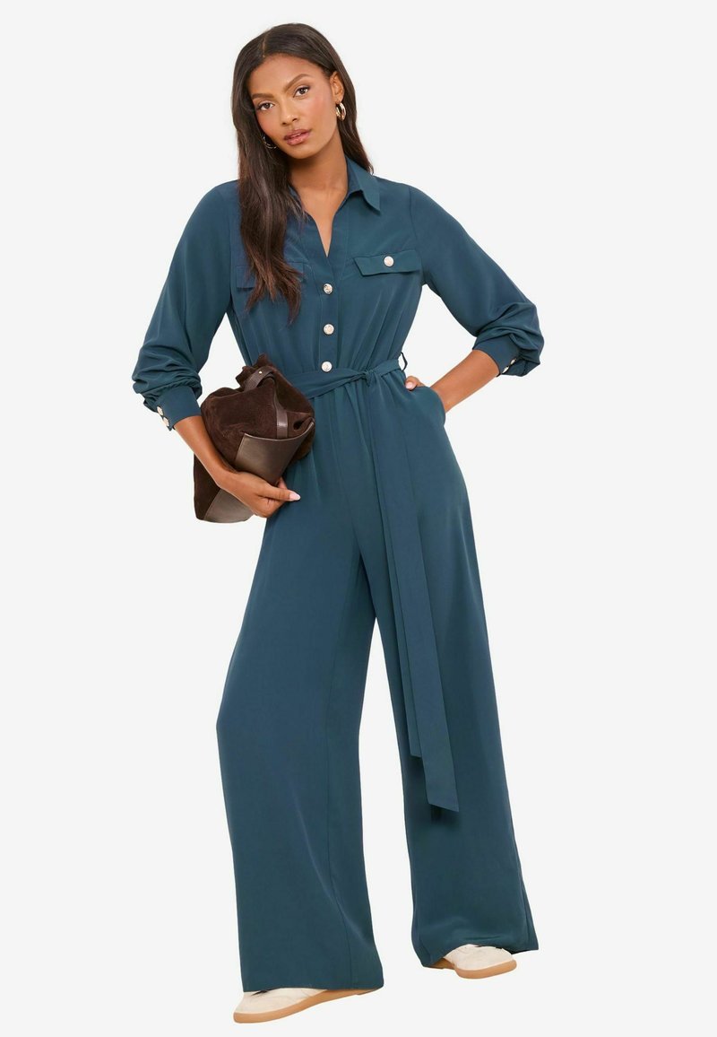 Türkisfarbener Overall mit langen Ärmeln, Knopfdetails und einem Taillengürtel. Verfügt über weite Hosenbeine und einen Kragen. Das Model hält eine braune Tasche.