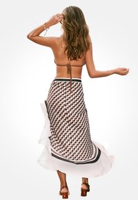 Mujer con el cabello largo, vestida con un top de bikini marrón, una falda midi con patrón geométrico y sandalias de tacón marrones, de pie de espaldas a la cámara.