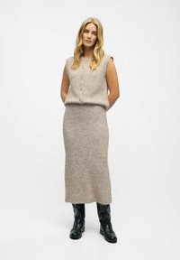 Ensemble en tricot deux pièces de couleur beige clair, comprenant un haut sans manches avec des boutons et une longue jupe. Porté avec des bottes noires à hauteur de genou.