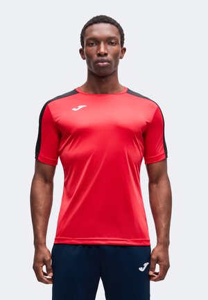 Homme portant un t-shirt de sport rouge avec des bandes noires aux épaules et un short bleu marine avec un logo blanc sur les deux vêtements, debout devant un fond uni.