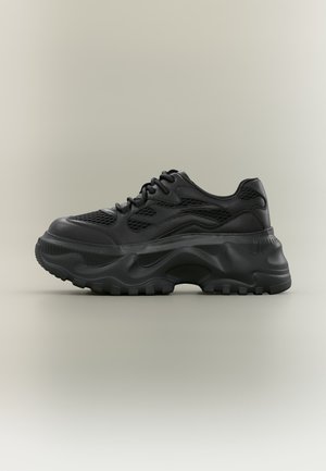 Zapatilla negra gruesa con suela gruesa y texturizada, parte superior de malla y cuero, y diseño con cordones sobre un fondo neutro.