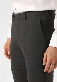 Pantaloni grigi su misura con fronte piatto, texture fine e passanti per la cintura. Abbinati a una camicia bianca con bottoni. Primo piano della vita e della mano.