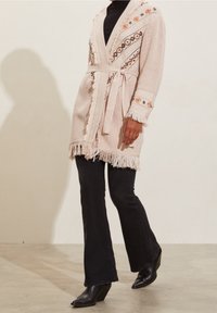Light pink cardigan with fringe hem and detailed embroidery, featuring a belted waist and a relaxed fit, paired with black flared jeans and heels.