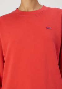 Sudadera de algodón roja con cuello redondo, que presenta un pequeño parche con el logo de Levis y una textura suave y lisa. Ajuste estándar y mangas largas.