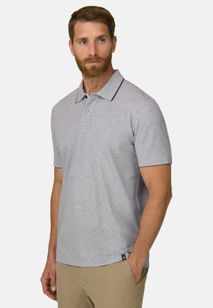 Mand med skæg iført lysegrå kortærmet poloshirt og beige bukser, stående med den ene hånd i lommen og kigger til siden.