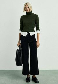 Maglione verde a collo alto indossato sopra una camicia bianca, abbinato a pantaloni neri a gamba larga. Una borsa a tracolla nera e mocassini completano il look.