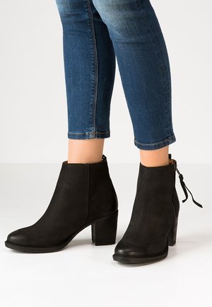 Zwarte suède enkellaarsjes met blokhakken en zijrits, gedragen bij cropped blauwe jeans tegen een effen witte achtergrond.