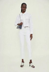 White button-up shirt with structured design, paired with white slim-fit pants. Model wears silver heeled sandals and holds a small gray bag.