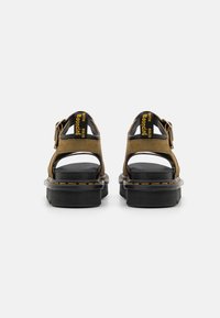 Sandalias de ante marrón con suelas negras. Parte trasera abierta, hebilla ajustable, texto amarillo "Bouncing" y costura amarilla a lo largo del borde.