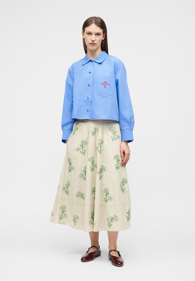 Γυναίκα που φοράει μπλε πουκάμισο με κουμπιά με το κείμενο «Yacht Club» και εκρού φούστα με πράσινα λουλουδάτα σχέδια, συνδυασμένα με σκούρα καφέ παπούτσια Mary Jane.