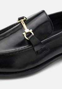 Mocassins en cuir noir avec une finition brillante, dotés d'un accent en métal doré et d'une bandoulière décorative. Design élégant à bout rond.
